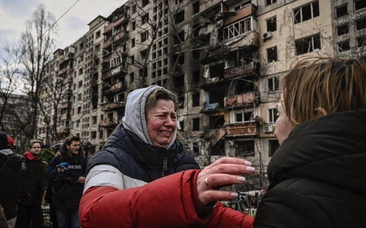 Kobieta w emocjonalnym uścisku z inną osobą, w tle zniszczony budynek.