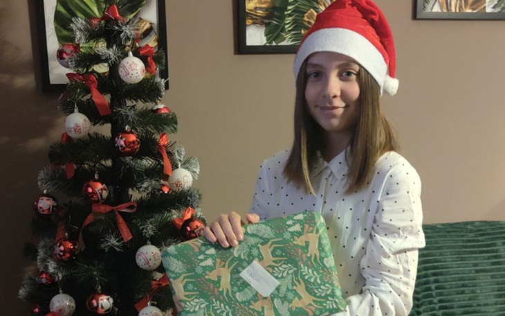 Dziewczyna w czapce Mikołaja, trzymająca prezent przy choince.