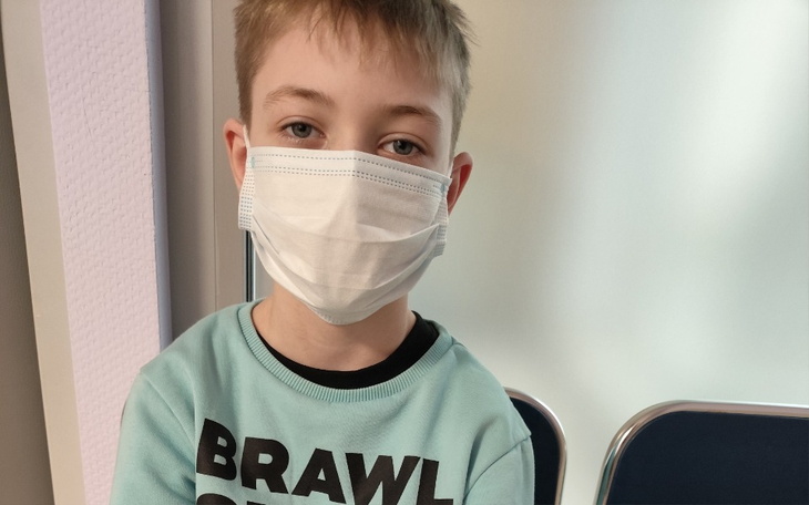 Chłopiec w maseczce, siedzący na krześle, z poważnym wyrazem twarzy.