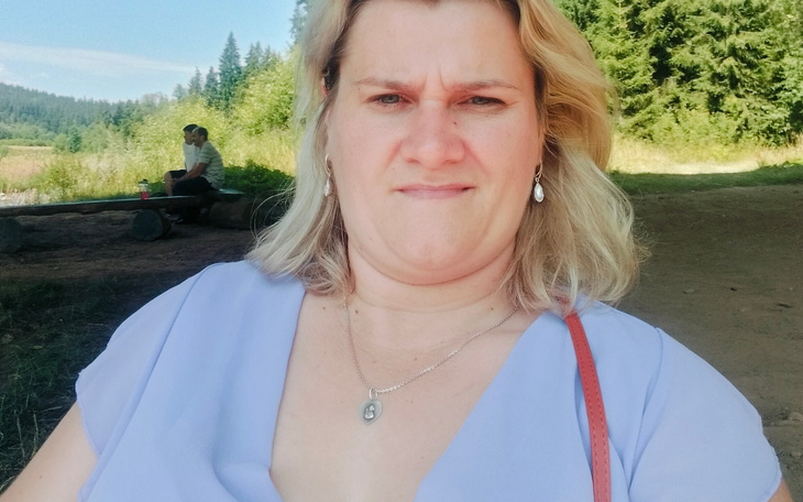 Selfie kobiety na zewnątrz, zielone drzewa w tle, uśmiech i srebrne kolczyki.