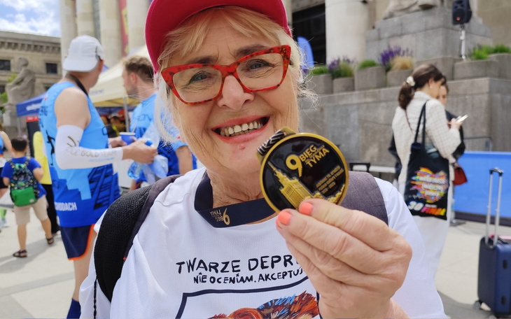 Uśmiechnięta kobieta trzyma medal po biegu na tle budynków.