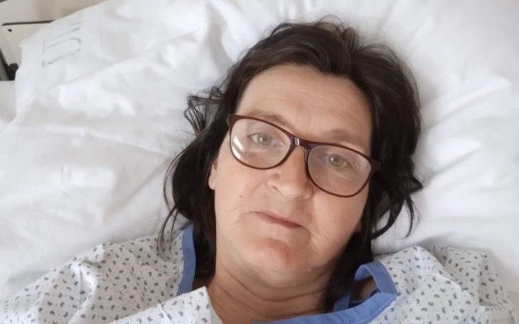 Kobieta w szpitalu, leżąca na łóżku, z okularami i spokojnym wyrazem twarzy.