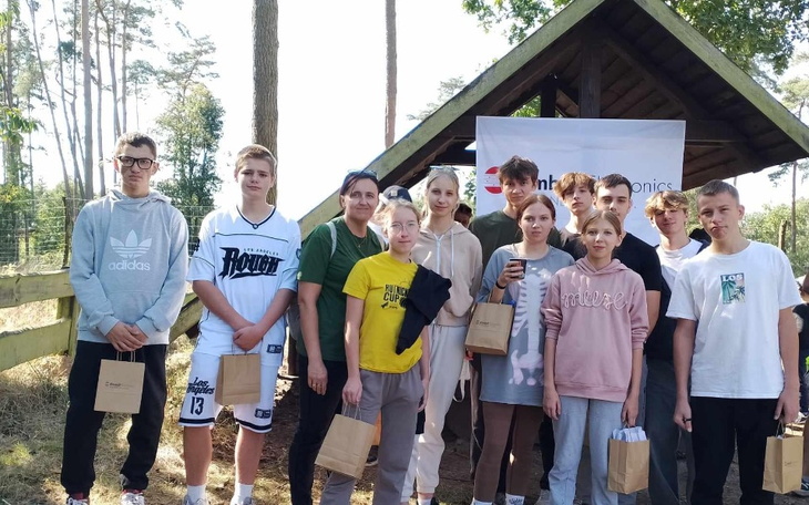 Grupa młodzieży z opiekunem na tle leśnej chaty, wszyscy uśmiechnięci.