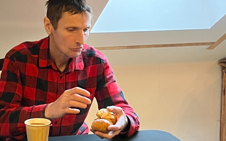 Mężczyzna w koszuli w kratę trzymający bułkę i filiżankę.