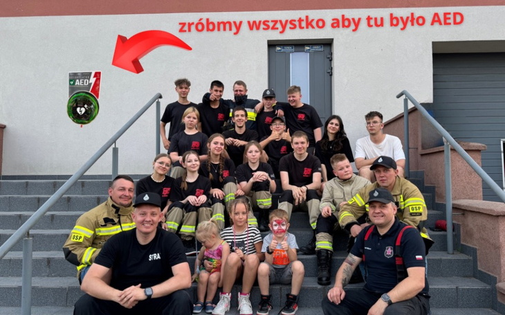 Grupa ludzi pozująca do zdjęcia przed budynkiem, promująca AED.