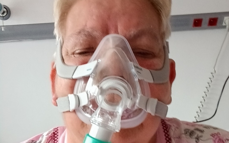 Kobieta w szpitalu, uśmiechająca się przez maskę tlenową.