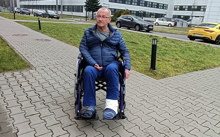 Starszy mężczyzna w wózku inwalidzkim z opatrunkiem na nodze, siedzący na chodniku.