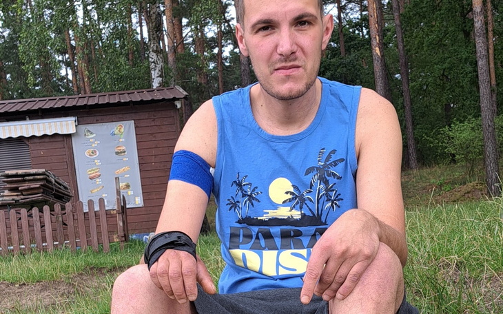 Mężczyzna w niebieskiej koszulce, siedzący na trawie w lesie.