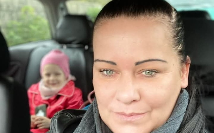 Kobieta w samochodzie, z dzieckiem w tle, obie uśmiechnięte.