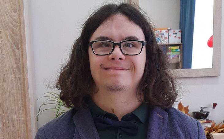 Mężczyzna z długimi włosami i okularami, uśmiechający się w eleganckim stroju.