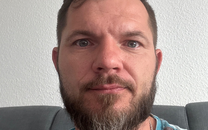 Mężczyzna z brodą siedzący na kanapie, patrzący w kamerę.