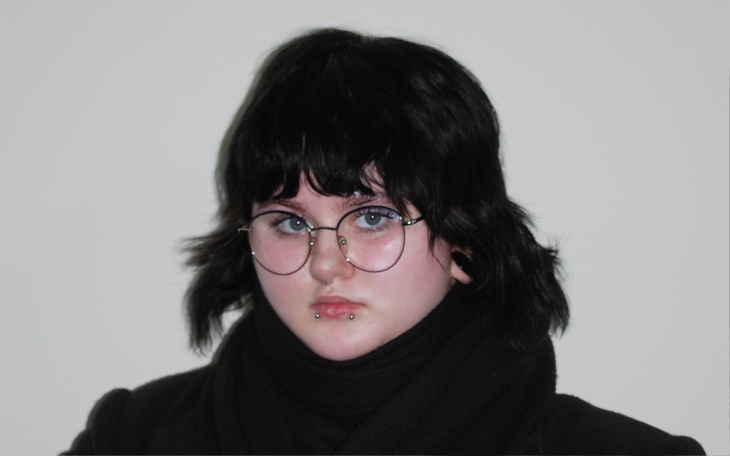 Młoda osoba z okularami i ciemnymi włosami na jasnym tle.