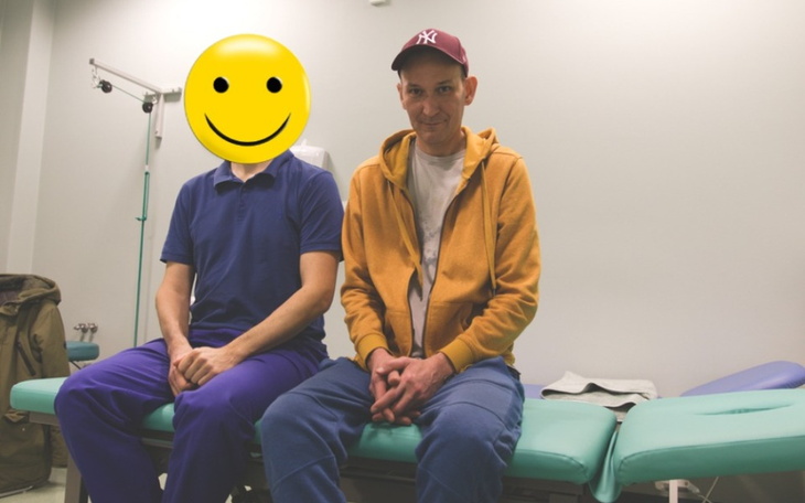 Dwóch mężczyzn siedzących na łóżku szpitalnym, uśmiechnięci.