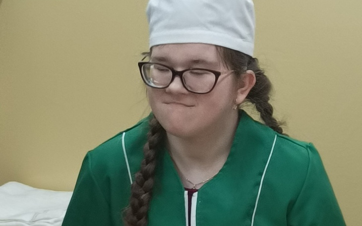 Młoda dziewczyna w zielonym fartuchu i czapce kucharskiej, z warkoczami.