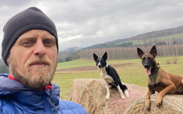 Mężczyzna z długimi włosami, pozujący z psem na tle krajobrazu.