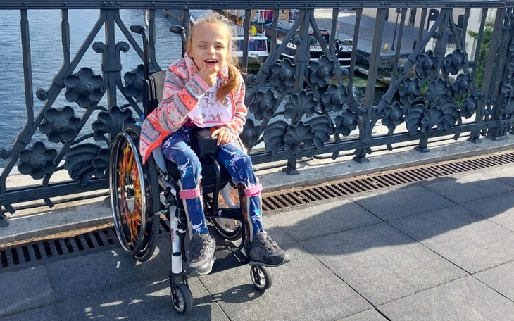 Dziewczynka na wózku inwalidzkim, uśmiechająca się na tle mostu.
