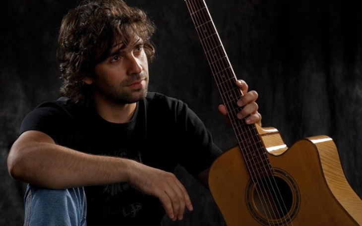 Mężczyzna z gitarą, w zamyślonej pozie, w ciemnym otoczeniu.