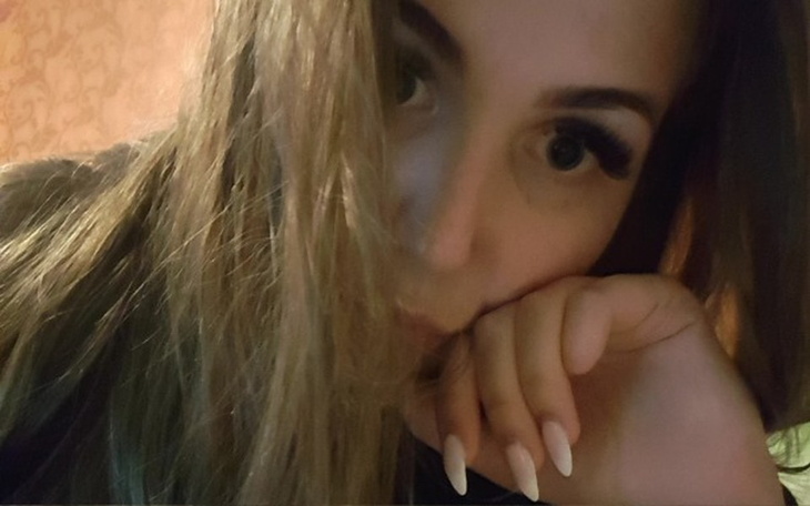 Osoba z długimi włosami, patrząca w obiektyw z ręką przy twarzy.