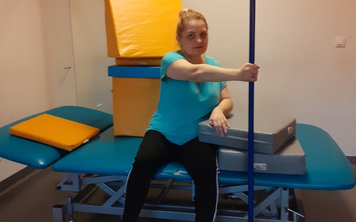 Kobieta siedząca na stole rehabilitacyjnym, otoczona poduszkami.