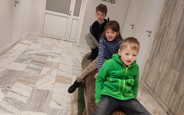 Troje dzieci bawiących się na dużym kłodzie w jasnym korytarzu.