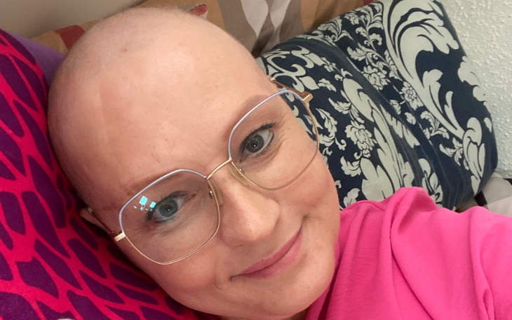 Kobieta z okularami i różową bluzą, leżąca na poduszkach, z uśmiechem na twarzy.