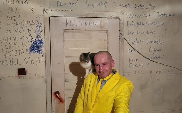 Mężczyzna w żółtej kurtce z kotem na ramieniu, pozujący przed drzwiami.