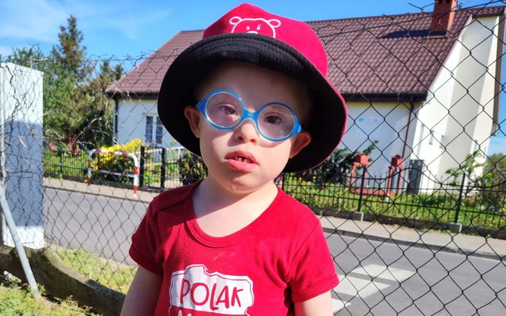 Chłopiec w kapeluszu i okularach, uśmiechający się na tle domu.