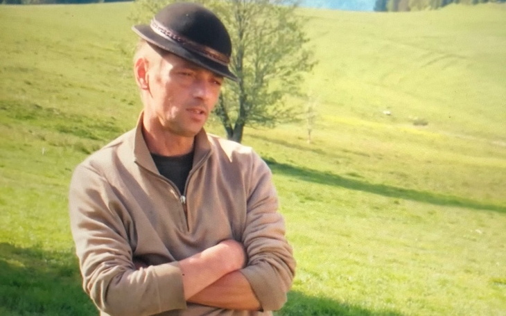 Mężczyzna w swetrze, z rękami skrzyżowanymi, na tle zieleni.