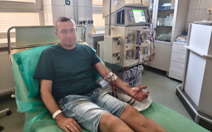 Chory mężczyzna w szpitalnym łóżku, na sali z aparaturą dializacyjną w tle.