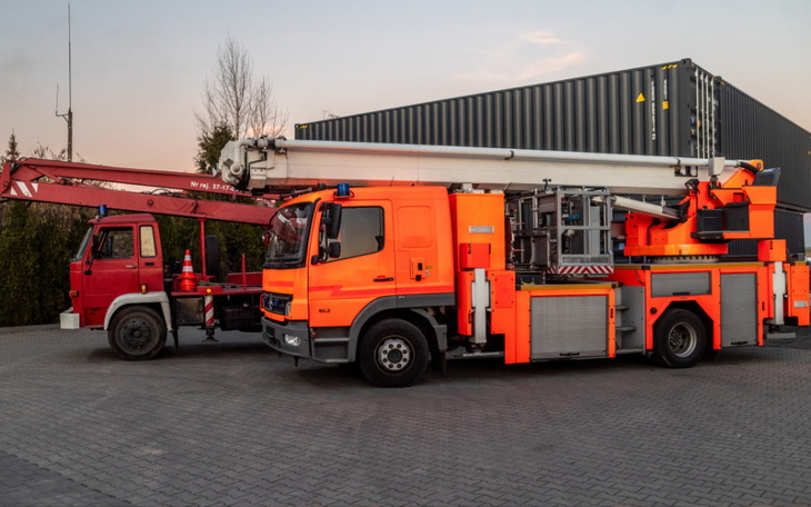 Pomarańczowy samochód strażacki z podnośnikiem, na tle budynku.