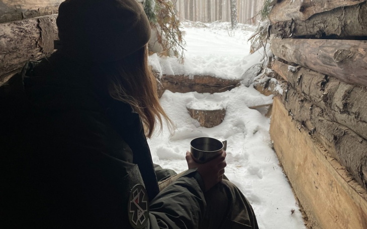 Osoba siedząca w schronieniu, trzymająca kubek w zimowym lesie.