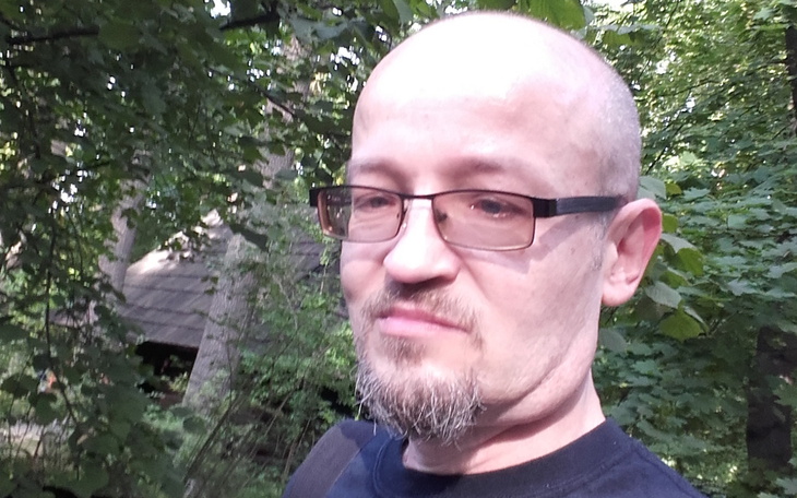 Mężczyzna z okularami w lesie, z drzewami w tle.