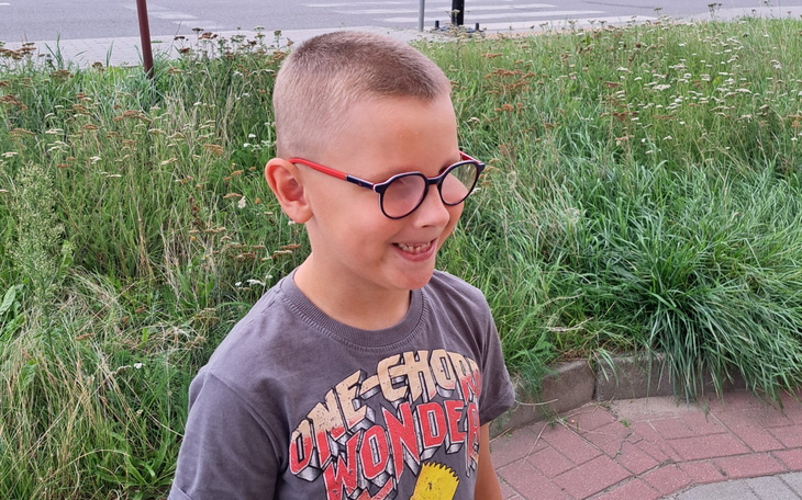 Uśmiechnięty chłopiec z okularami w terenowej scenerii z trawami i kwiatami.