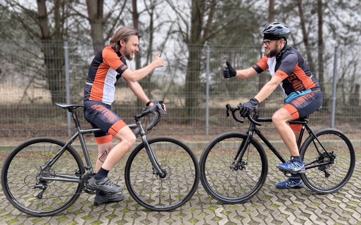 Dwaj rowerzyści w strojach sportowych, uśmiechają się i pokazują kciuki w górę.