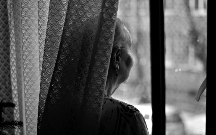 Starsza kobieta patrzy przez zasłonę, zamyślona i refleksyjna.