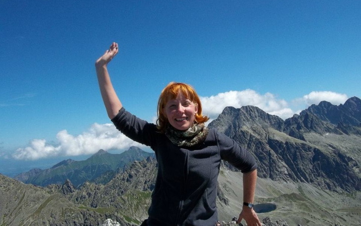 Kobieta na szczycie góry, machająca ręką w radosnym geście.