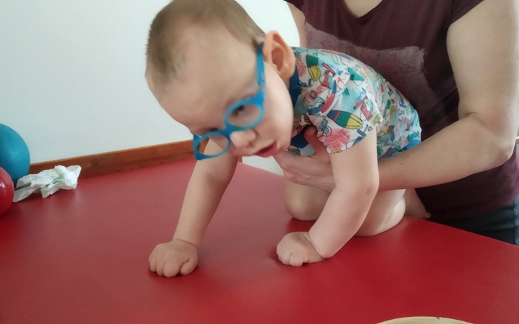 Małe dziecko w okularach, wspierane przez dorosłego na stole.