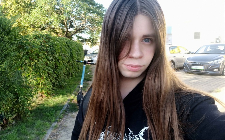 Młoda osoba z długimi włosami, selfie na tle zieleni.