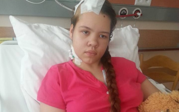 Młoda dziewczyna w szpitalu, z opatrunkiem na głowie, trzyma pluszowego misia.