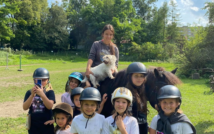 Grupa dzieci w kaskach, z kobietą na koniu w tle.