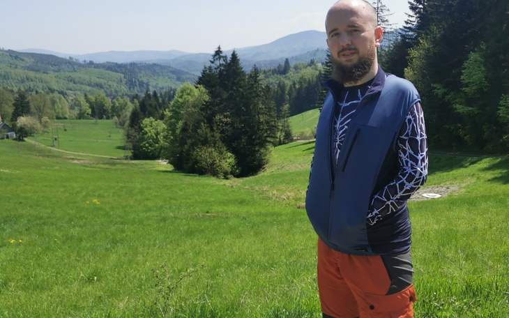 Mężczyzna w kolorowej odzieży stoi na zielonym wzgórzu, w tle las.