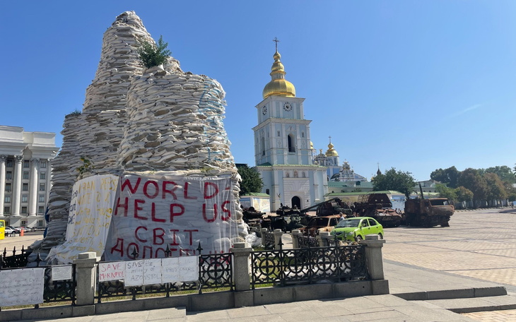 Pomnik z napisem 'WORLD HELP US' w tle z cerkwią.