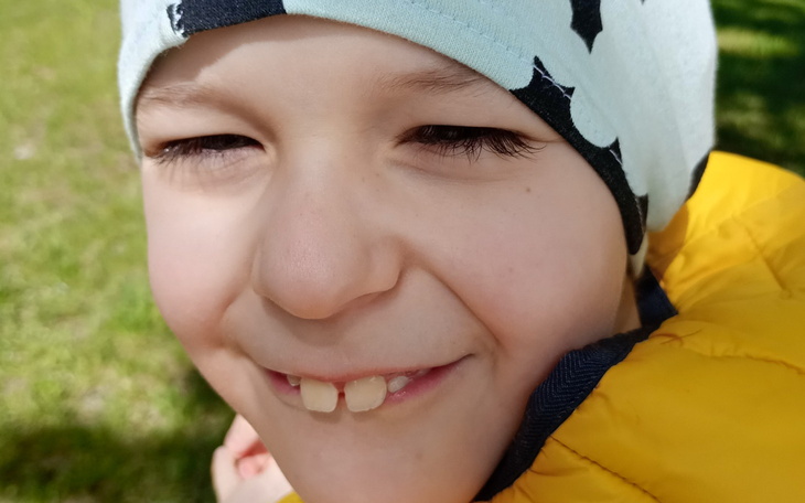 Uśmiechnięty chłopiec w żółtej kurtce i czapce na tle zielonej trawy.