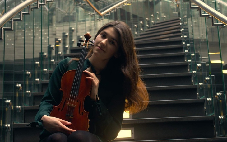 Młoda kobieta z wiolonczelą, siedząca na schodach w nowoczesnym wnętrzu.