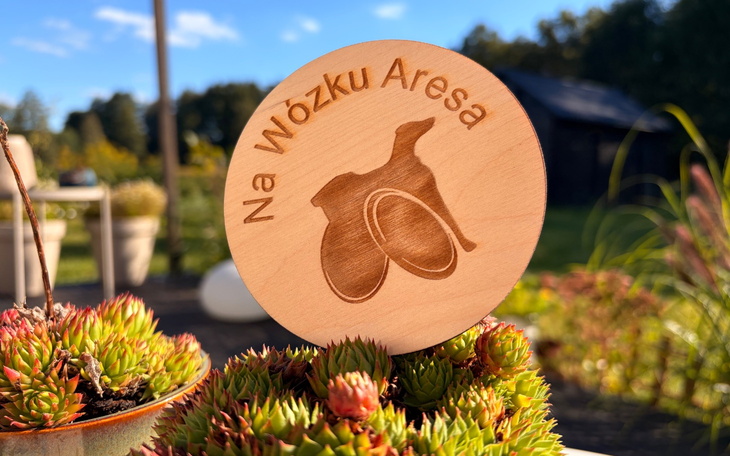 Drewniana tabliczka z napisem 'Na Wózku Aresa' na tle roślin.