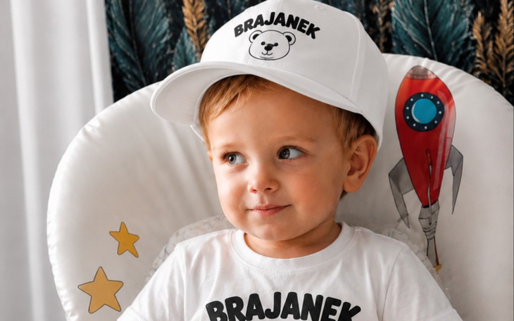 Uśmiechnięte dziecko w białej czapce z napisem BRAJANEK
