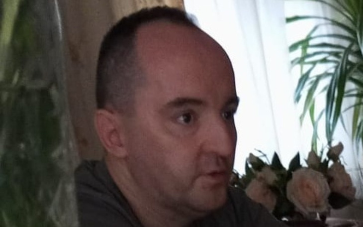 Mężczyzna w szarej koszulce, siedzący przy stole z kwiatami w tle.