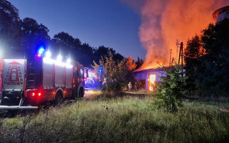 Straż pożarna walcząca z pożarem w nocy, w tle płonący budynek.