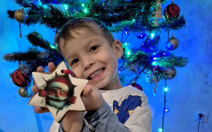 Chłopiec trzyma ozdobę świąteczną przed choinką z kolorowymi światełkami.