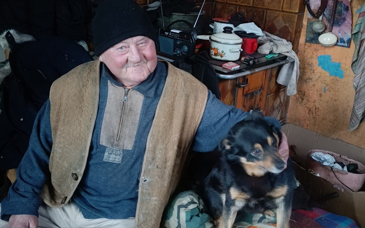 Starszy mężczyzna z uśmiechem siedzi z psem w skromnym wnętrzu.